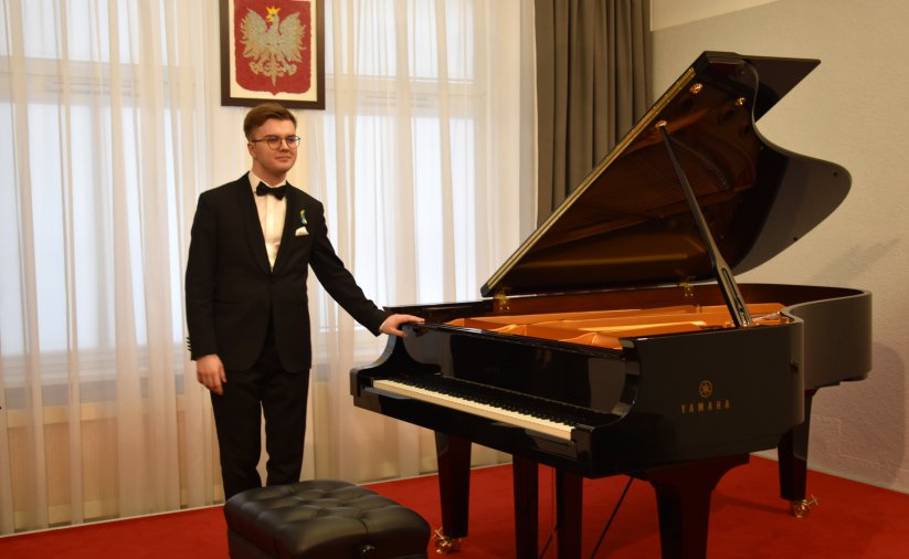 Artysta stojący przy fortepianie, w drugim planie godło Polski