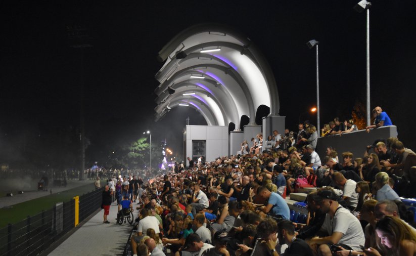 Trybuna stadionu wieczorem pełna ludzi, nad częścią trybuny zadaszenie