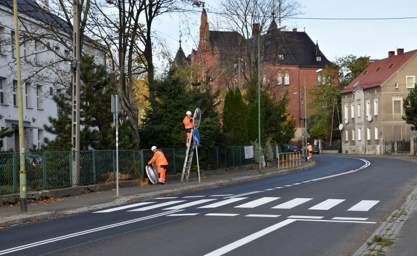 Przejście dla pieszych, dwie osoby w pomarańczowych kamizelkach montują znak drogowy
