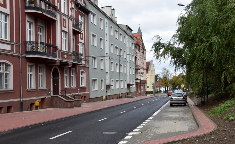 Dwie staromiejskie kamienice, droga i zaparkowane obok auta, po prawej zieleń drzew