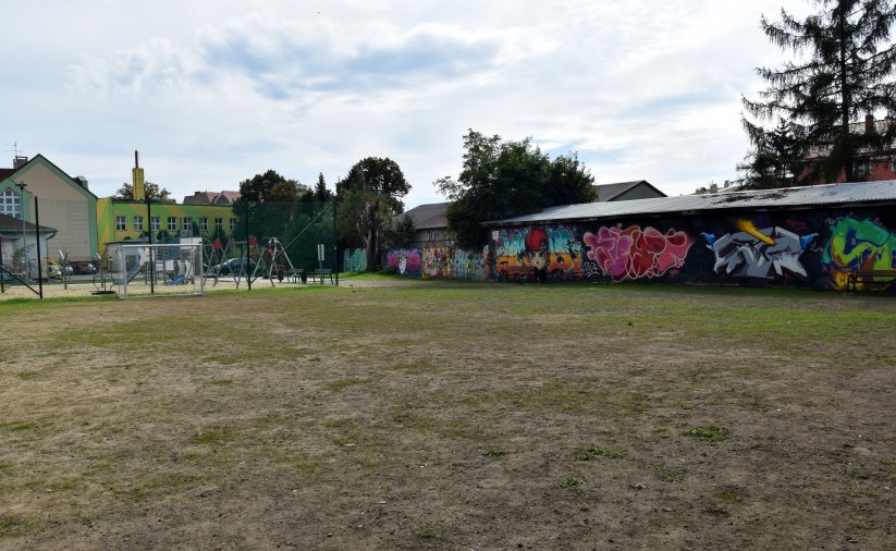 Boisko trawiaste, obok długi parterowy budynek z namalowanym kolorowym grafitti