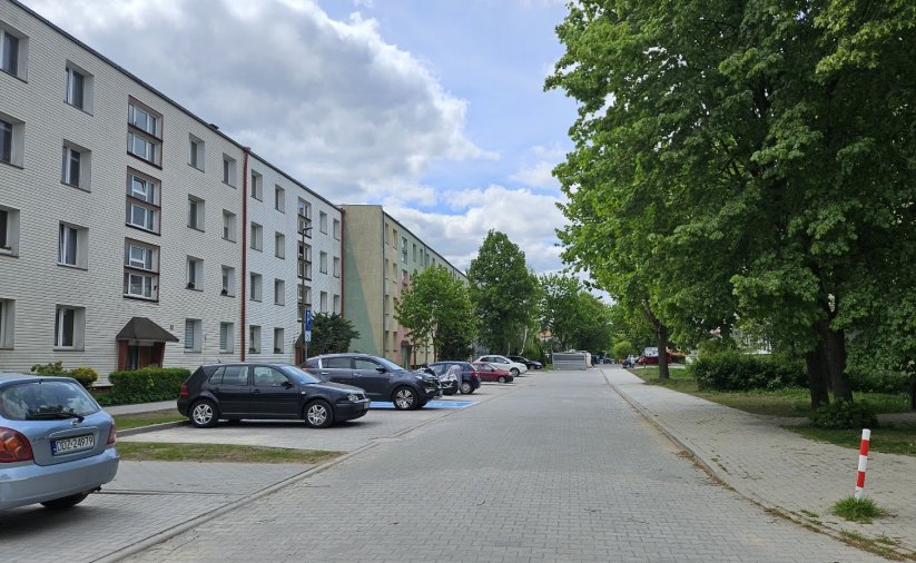 Droga osiedlowa z kostki betonowej, po lewej blok, po prawej duże drzewa