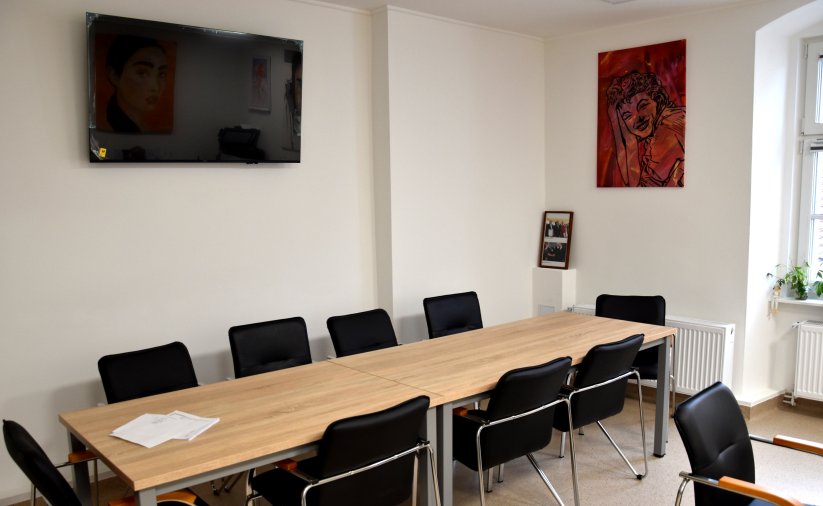 Stół i czarne krzesła, na białych ścianach obraz i telewizor