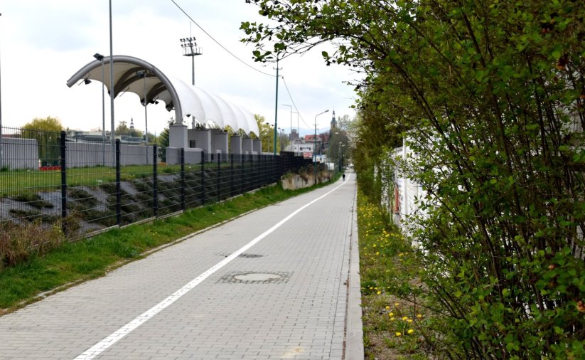 Bardzo długi chodnik z kostki betonowej, po prawej zieleń, po lewej trybumy stadionu