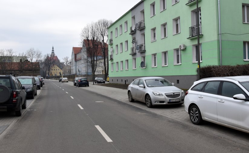 Droga asfaltowa, po bokach chodniki z zaparkowanymi autami, po prawej zielony trzypiętrowy blok