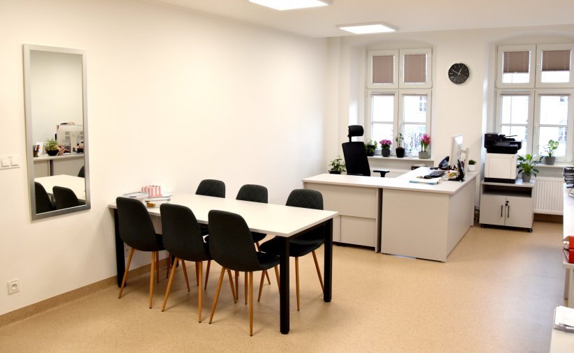 Duże jasne pomieszczenie, stół ustawiony przy ścianie, przy oknach biurko