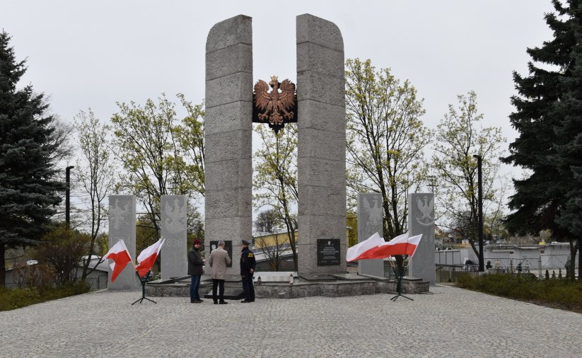 Pomnik, przed nim flagi, po bokach drzewa iglaste