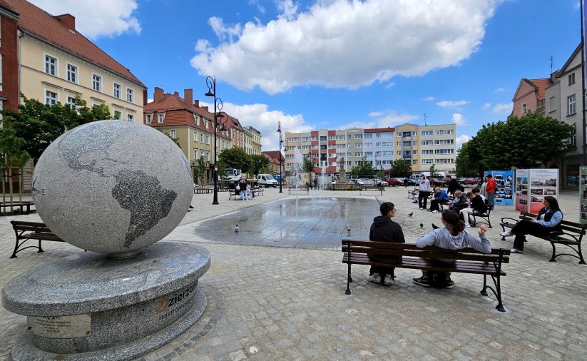 Duży kamienny globus ustawiony w rynku, obok ławka, na drugim planie fontanna
