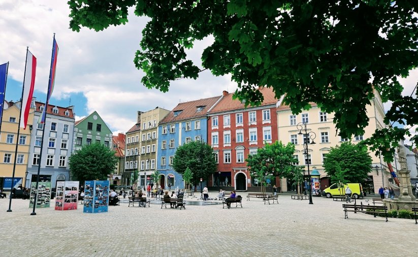 Kamienice w rynku, po prawej zieleń po lewej maszty z pionowymi flagami