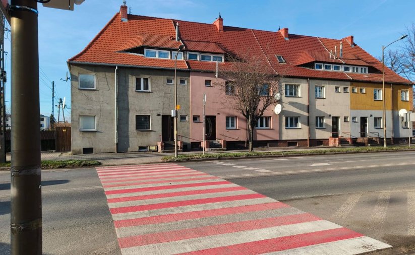 Przejście dla pieszych w białe i czerwone pasy, za nim długa kamienica