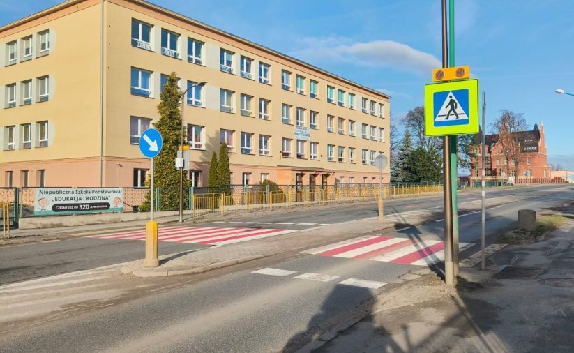 Szeroka droga, białoczerwone przejścia dla pieszych i duży czteropiętrowy żółty budynek