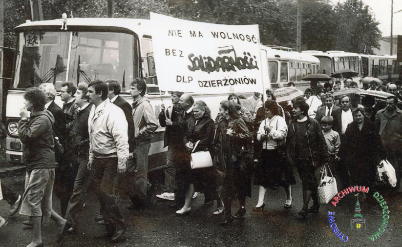 Czarno biała fotografia grupy ludzi przy autobusie z transparentem solidarności