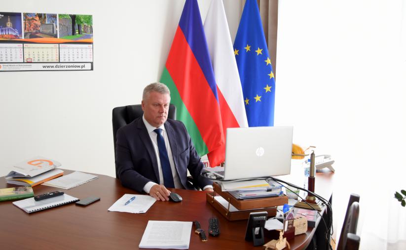 Burmistrz siedzący przy biurku, w tle flagi Polski, UE i Dzierżoniowa