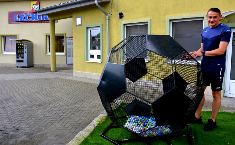 Kosz na nakręktu w kształcie czarnej piłki, obo stojący mężczyzna,w drugim tle żólty budynek