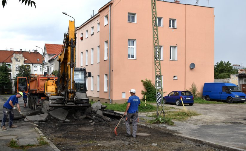 Koparka pracująća na remontowanej drodze, przy koparce pracownik, w tle budynek