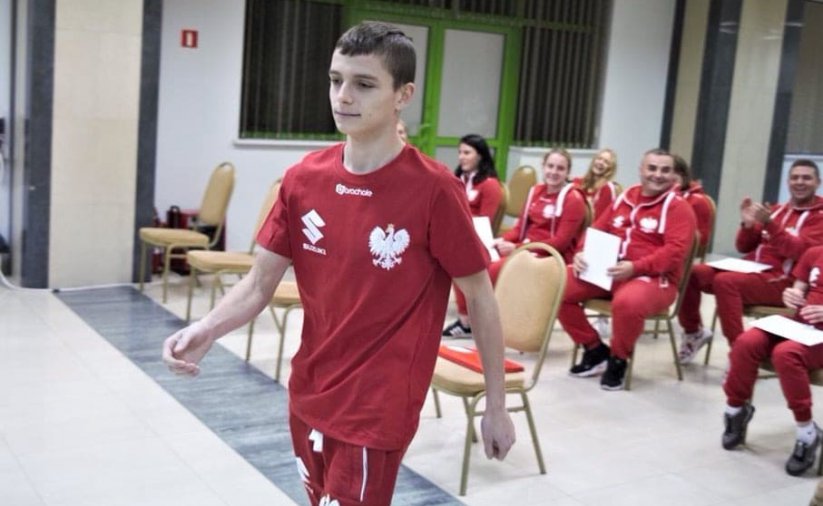 Młody zawodnik idący w koszulce z godłem, w tle gupa siedzących osób