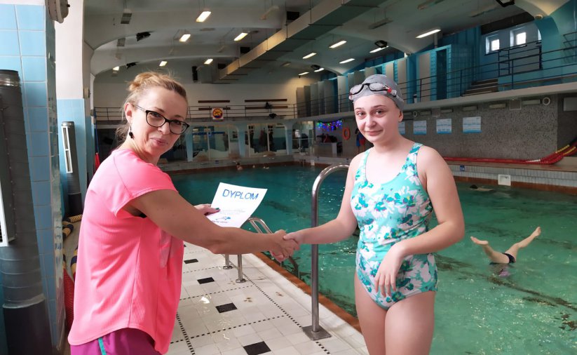Na zdjęciu intruktor pływania wręcza dyplom dziewczynce, podają sobie dłonie, stoją przy basenie