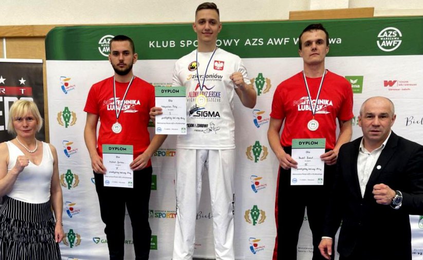 Trójka zawodników stojąca na podium, obok podium dwie osoby