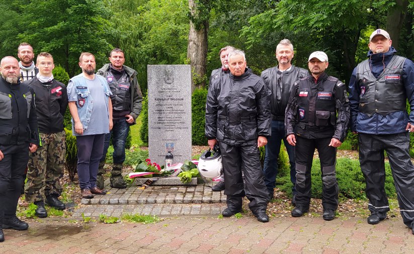 Grupa mężczyzn w strojach motocyklowych stojąca przy kamiennym obelisku 