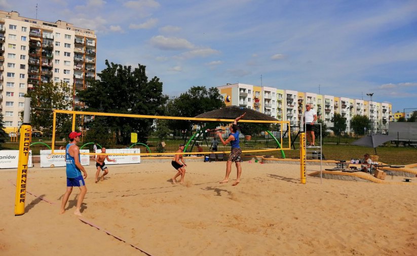Cztery osoby grają w siatkówke plażową, w drugim tle osiedle mieszkaniowe