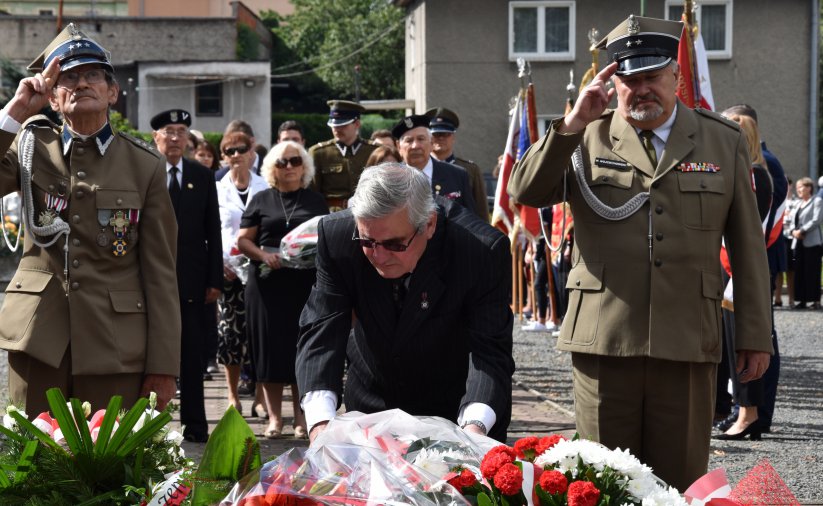 Kombatanci salutujący przy pomniku, na pierwszym planie wieńce kwiatów