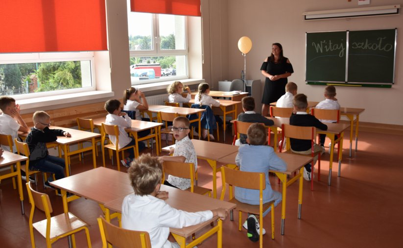 Szkolna klasa, uczniowie siedzący przy łąwkach i uśmiechnięty nauczyciel