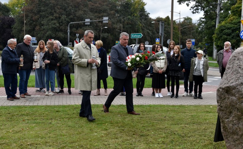 Grupa stojących osób, dwóch panów ze zniczem i kwiatami idą w kierunku kamiennego obelisku