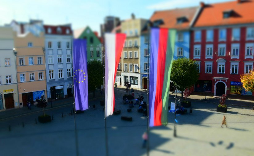 Maszty z flagami Polski, UE i Dzierżoniowa, z tyłu zabytkowe kamienice