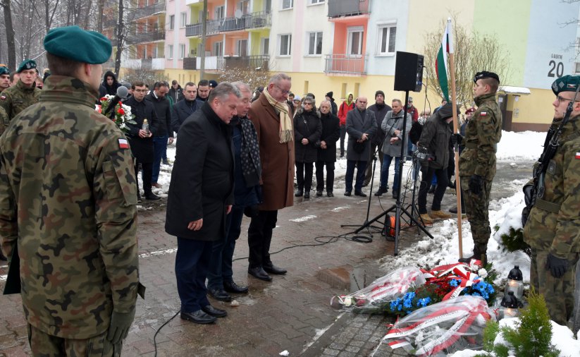 Liczna grupa osób, wśród nich żołnieże, trzy osoby składają ukłon przed obeliskiem