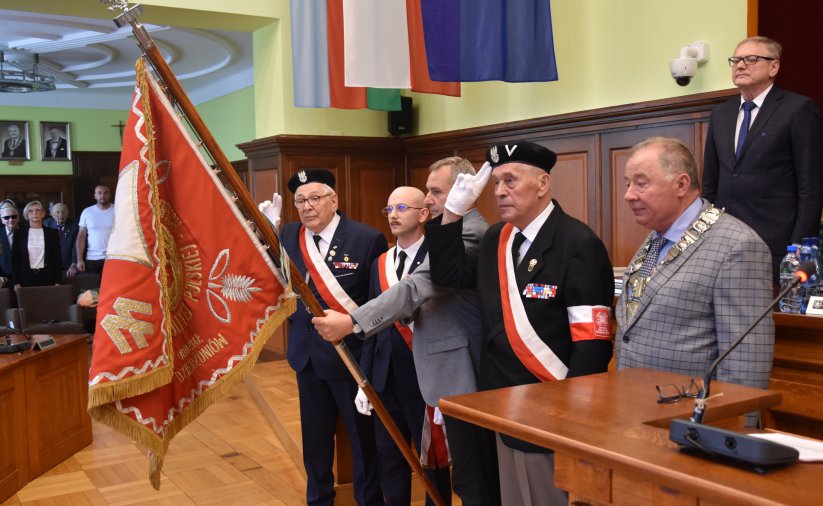 Kombatanci w mundurach trzymają czerwony sztandar i salutują