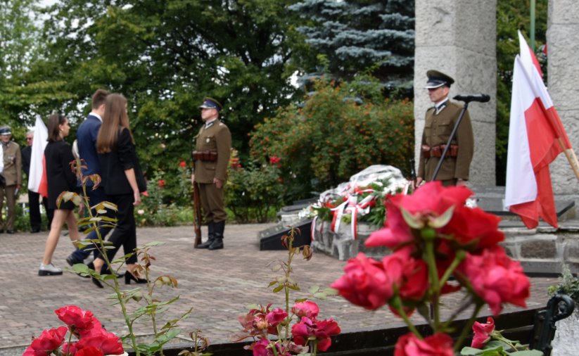 Dwóch żołnieży pełni wartę przy pomniku, do któego podchodzi grupa młodzieży z kwiatami, obok flaga Polski