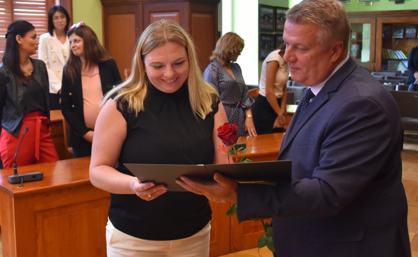 Uśmiechnięta kobiet w czarnej bluzce odbiera list gratulacyjny o mężczyzny w garniturze, w tle grupa kobiet