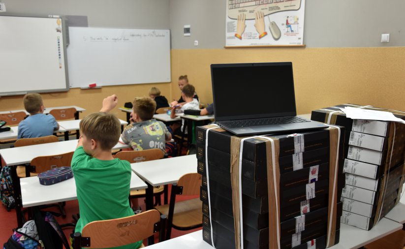 Uczniowie podczas zajęć w klasie, na pierwszym planie laptop i uczeń z podniesioną ręką 