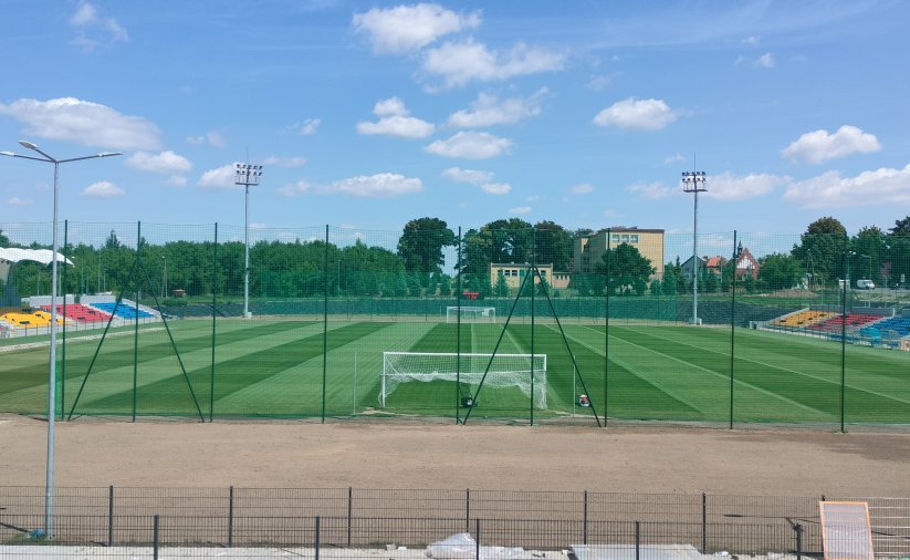 Stadio, widok z góry od strony bramki. Po bokach kolorowe trybuny, w tle zieleń drzew