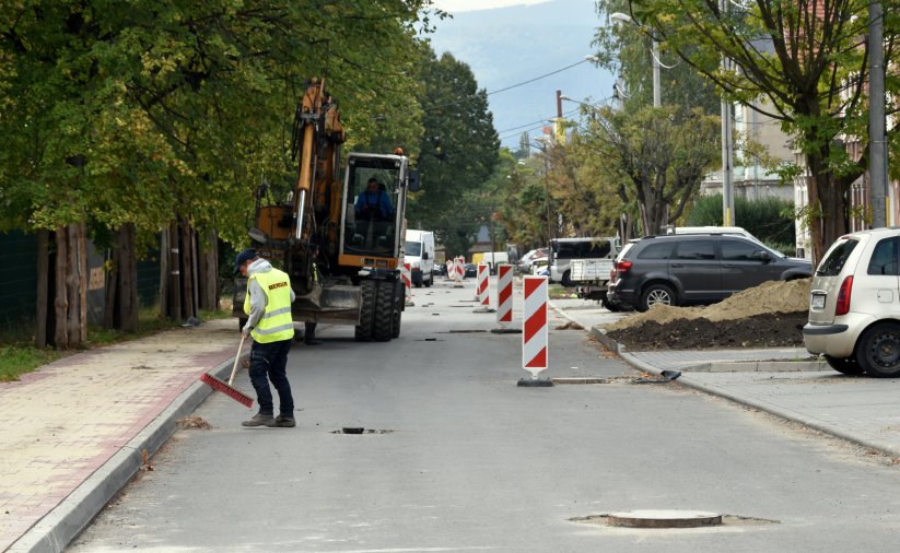 Prace przy budowie drogi, człowiek w jaskrawej kamizelce zamiata drogę, za nim koparka