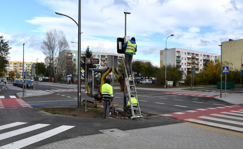 Trzy osby w jaskrawych kamizelkach montuje znak przy skrzywoniu dróg, w tle bloki mieszkalne