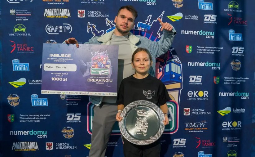 Trener trzyma w reku duży czek, przed nim zawodniczka ze srebrną paterą w tle ścianka promocyjna