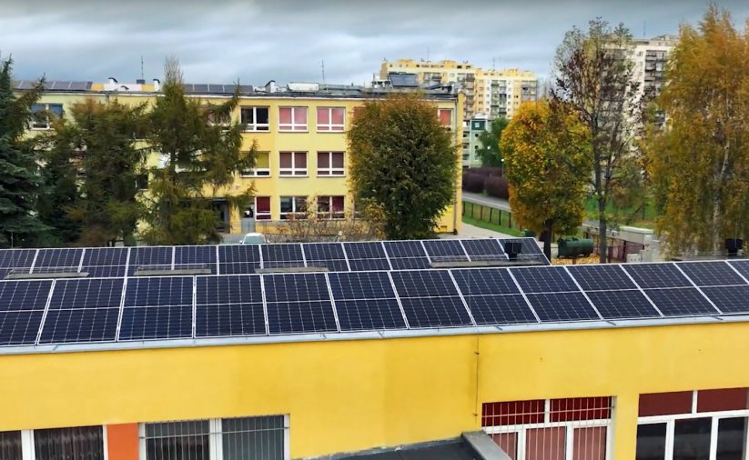 Panele fotowoltaiczne na dachu budynku, w tle osiedle mieszkaniowe