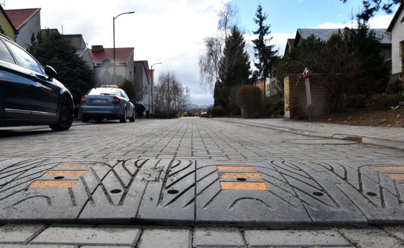 Droga z kostki betonowej i widoczny na niej "spowalniacz", o lewej dwa zaparkowane auta