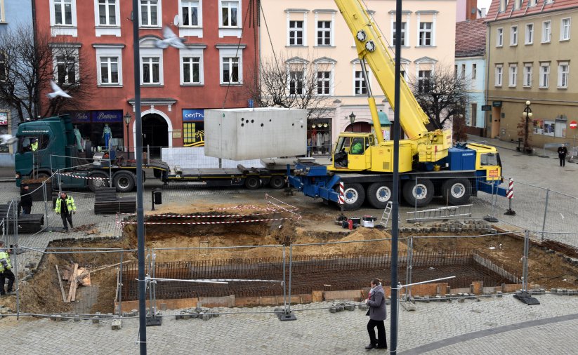 Dźwig unoszący dużą betonową konstrukcję, w tle kamienice