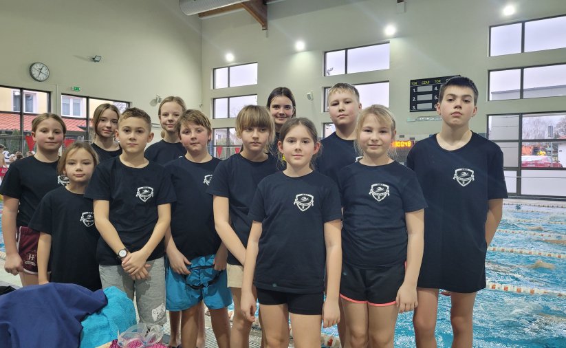 Grupa młodych pływaków i pływaczek w ciemnych koszulkach, za nimi basen