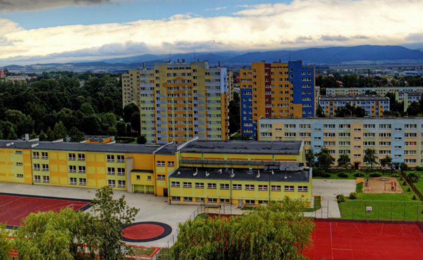 Budynek szkoły z góry, przed nim dwa czerwone boiska sportowe, za budynkiem bloki i wieżowce oseidlowe, w dalekim planie góry