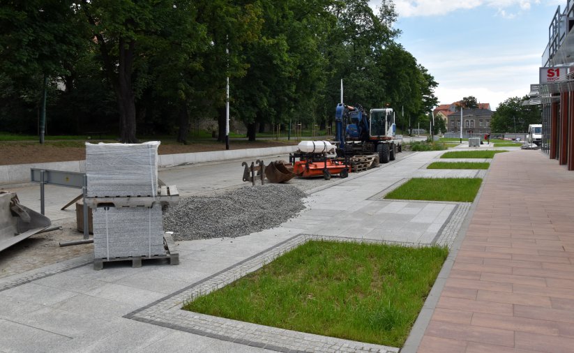 Droga z kostki kamiennej w budowie, obok małe skwery zieleni, na drodze maszyny budowlane