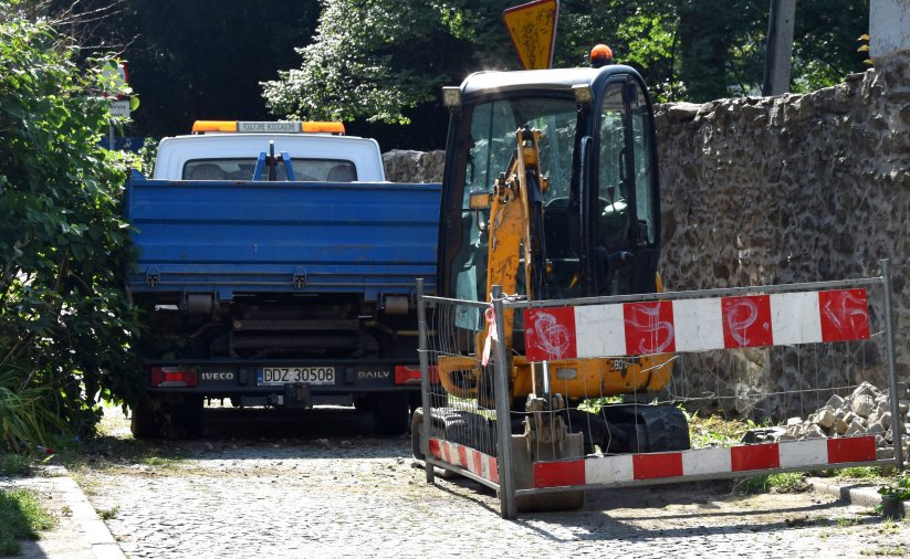 Mała koparka i mały samochód dostawczy na wąskiej drodze, obok drogi kamienny mur