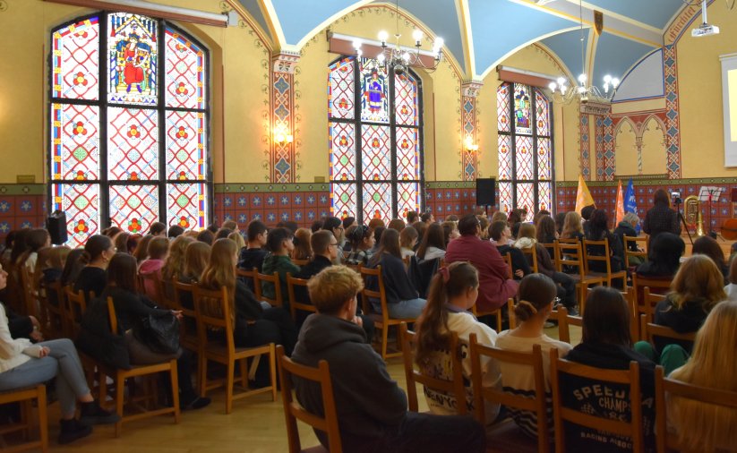 Aula szkolna z dużymi okiennymi witrażami zapełniona uczniami