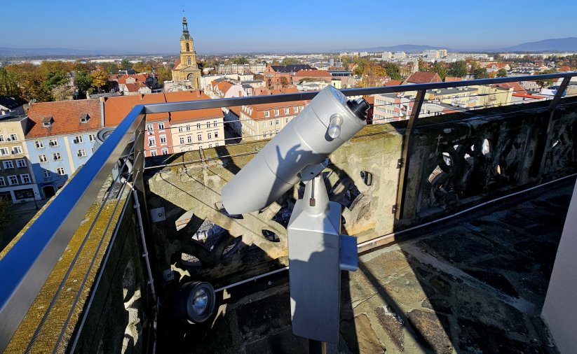 Wieża ratuszowa z kaminnymi i metalowymi barierkami, na pierwszym planie luneta, w tle panorama miasta