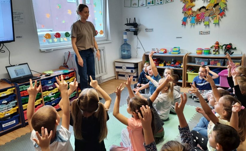Przedszkolaki siedzący w grupie zgłaszają się do odpowiedzi, przed nimi młoda kobieta