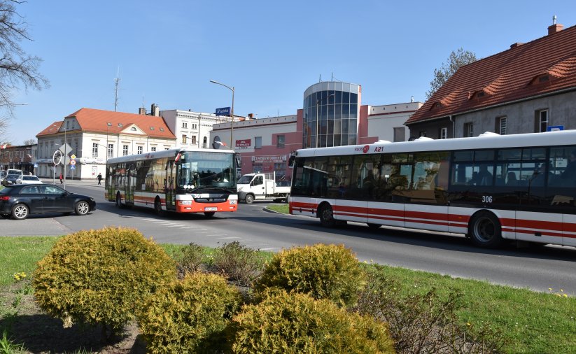 Dwa mijające się na drodze autobusy komunikacji miejskiej, z przodu zieleń, w tle budynki