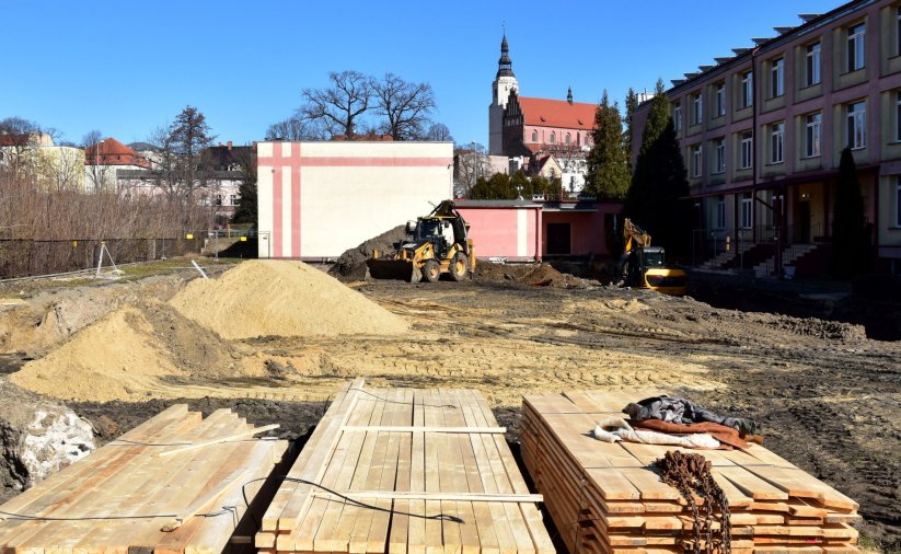 Plac budowy z materiałami budowlanymi, obok trzypiętrowy budynek szkoły