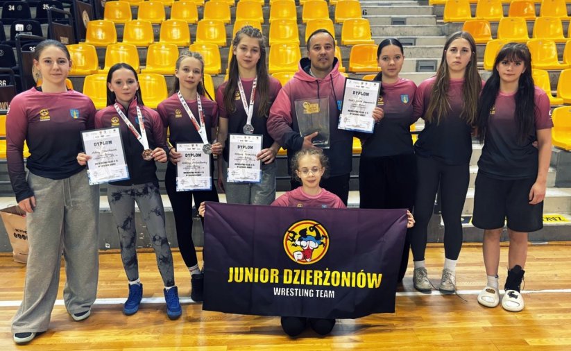 Zapaśniczki w jednolitych strojach z dyplomami z rekach, po środku trener